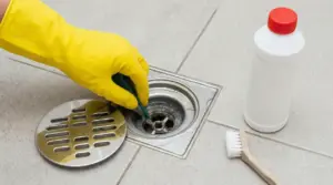 Mano con guanto giallo pulisce lo scarico della doccia con spugna e detergente sul pavimento del bagno